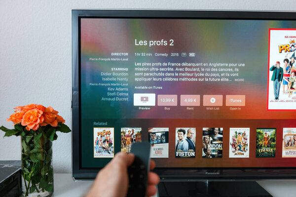 Paris, France - Nov 25, 2015: A male hand points the remote control of Apple TV towards a plasma 4K TV in the living room, with less glare on the screen, while a vase of roses sits nearby