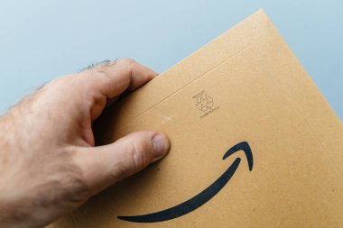 Paris, Frnace - 25 Ekim 2023: Bir erkek, teslim için hazırlanmış Amazon Prime karton paketini kavrıyor. Tanınabilir paketleme saf mavi bir arkaplan karşısında canlı bir tezat oluşturur.