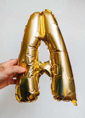 Parlak altın helyum balonlarından yapılmış A harfini, el değmemiş beyaz bir arka plana karşı tutan bir erkek el tutuyor.