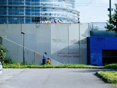 Strasbourg, Fransa - 8 Mayıs 2023: Yürümekte olan çocuğuyla bekar bir anne, protesto tabelalarının duvarları süslediği parlamento binasının önünden geçiyor. Günlük hayatın yan yana gelişine tanık olun.
