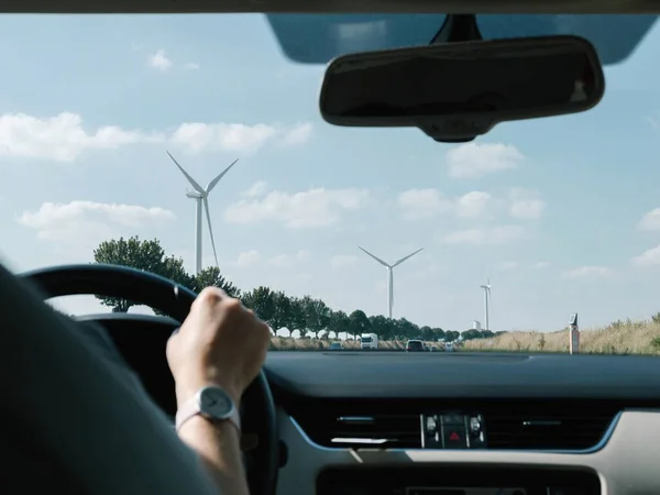Female hand holding steering wheel with multiple windmills in front of the driving road perspective
