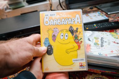 Paris, Fransa - 16 Aralık 2023: Aile eğlencesi için bir çocuğun ayaklarının yanında, bir televizyon setinin önünde Les Aventures de Barbapapa DVD 'sini tutan bir erkek..