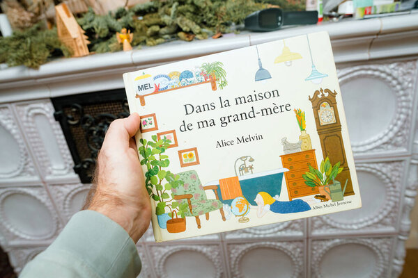 Paris, France - Dec 16, 2023: Male hand presenting Dans la maison de ma grand-mere by Alice Melvin, a kids book, in front of a rustic stove