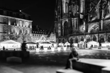 Kadınlar Majestic Place de Chateau 'ya karşı, Notre-Dame Katedralleri Noel Pazarı' nda alacakaranlıkta parlıyor. Oyuncak ve hediyelik eşya satan market tezgahları ve tezgahları olan siyah beyaz bir sahne.