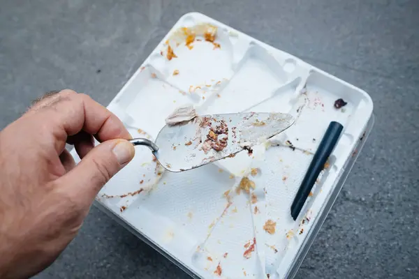 男性が壊れたスパチュラを保持し アフターパーティーの残骸の中で空のプラスチック製のケーキトレイの上に休むように侵入が展開されます — ストック写真