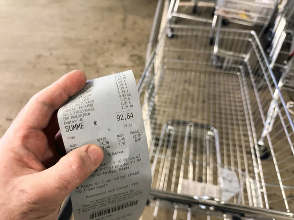 德国柏林 2024年1月23日 一只男性手拿着超市的一张纸收据 前面有一辆空购物车 总价签为92 64欧元 这说明了德语的概念 — 图库照片