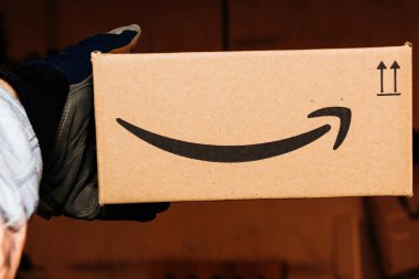 Bremen, Almanya - 10 Aralık 2023: Eldiven giyen bir adam, etkili ve güvenilir teslimat hizmetlerini sembolize eden gülümseyen ok logosuyla süslenmiş Amazon Prime karton paketini bir depoda tutuyor.