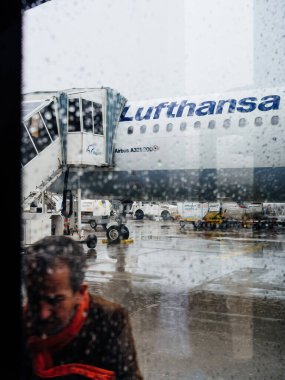 Frankfurt, germany - May 4, 2019: A person wearing a red scarf observed through a rain-speckled window against the backdrop of a Lufthansa Airbus A321. clipart