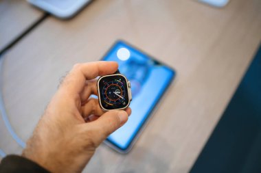 Paris, France - Oct 28, 2024:A male hand holds an Apple Watch Ultra, with a tablet visible in the defocused background, showcasing the smartwatchs detailed display and emphasizing the integration of clipart