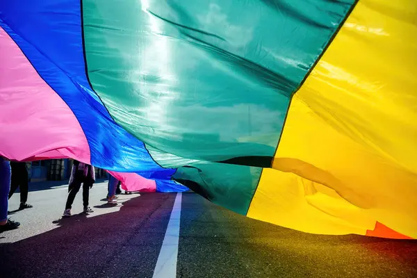 stock image People holding a vibrant rainbow flag at a pride parade, with sunlight casting beautiful shadows and highlighting the fabrics colors on the pavement below