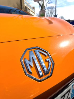 Freiburg, Almanya - Mar 2, 2024: Parlak elektrikli turuncu bir arabanın kaputundaki MG logosunun yakın görüntüsü. Canlı renk ve parlak amblem, bulanık şehir arkaplanı ve berrak