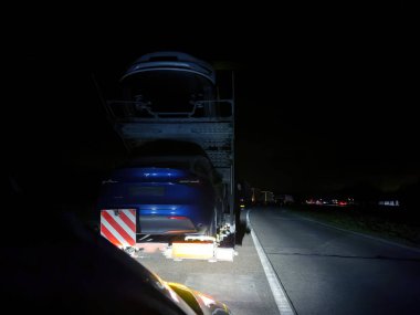 Freiburg, Almanya - 5 Mar 2024: Büyük bir trafik sıkışıklığı, bir römork üzerinde birden fazla yeni Tesla elektrikli araba taşıyan bir dizel kamyonu barındırır. Sahne, elektrikli araçlara geçişi simgeliyor.