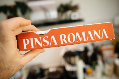 Erkek el, mutfak arka planında geleneksel İtalyan yemekleri olan lezzetli Pinsa Romana paketini tutuyor. Ev teslimatını ve aşçılık zevkini vurguluyor.