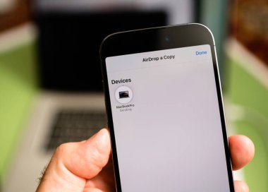 Paris, Fransa - Sep 29, 2022: Apple Bilgisayar 'ın Airdrop arayüzünü arka planda MacBook Pro ile gösteren bir iPhone' u tutan bir el, kablosuz olarak bir dosya gönderme sürecini resmediyor