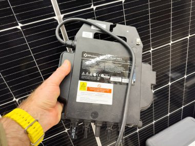 Paris, Fransa - 24 Nisan 2024: Sarı saat takan bir el bir güneş paneli dizisinin önünde bir APsystem DS3-L mikroçevirici tutuyor. Aygıt özellikleri ve güvenlik uyarıları açık