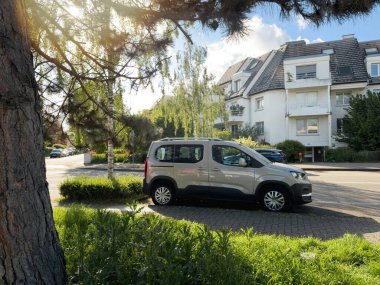 Paris, Fransa - 24 Nisan 2024: Güneşli bir iskan caddesinde park etmiş, yeşil ve ağaçlarla çevrili, arka planda modern bir apartmanı olan gümüş renkli bir Peugeot kargo arabası,