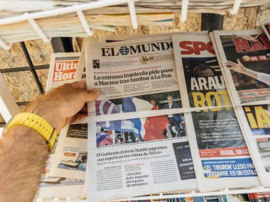 Valldemossa, İspanya - 8 Temmuz 2024: Sarı saati olan bir erkek el, İspanya 'nın Mallorca kentindeki bir basın standında Macron ve Afrikalı göçmenlerle ilgili manşetler atan El Mundo' nun bir sayısını elinde tutuyor..