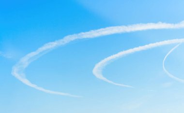 Beyaz buhar izleri, açık mavi gökyüzüne karşı kıvrımlı desenler oluşturur. Uçan uçakların karmaşık yollarını ve açık hava sahasının güzelliğini gözler önüne serer..