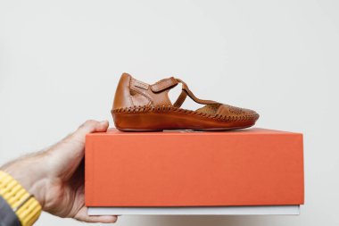 Turuncu bir ayakkabı kutusunda beyaz arka planda oturan kadınlar için örülmüş deri ayakkabılar tutan bir erkek el. Ürün, indirimli lüks bir ürün olarak konumlandırıldı..