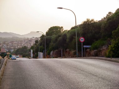 SEstany den Mas, Mallorca 'da yol manzarası, sokak lambaları ve 30 km hız sınırı tabelasıyla kaplı, günbatımında arka planda evleri ve tepeleri olan
