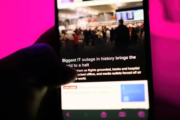 stock image Paris, France - Jul 20, 2024: A male hand holding an iPhone displays a news article about the biggest IT outage in history, which has brought the world to a halt, against a vibrant pink background