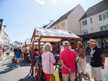 Hardelot, France - Aug 20, 2023:A vendor in a pink shirt engages in conversation with a customer at a beverage stall in the central market of Neufchatel-Hardelot, France, while passersby browse and clipart