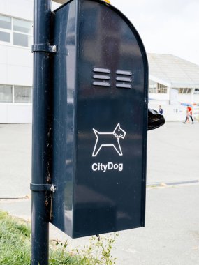 CityDog etiketli siyah bir çöp tenekesinde sokak yakınlarındaki bir kentsel alanda köpek atıklarının atılması için tasarlanmış bir köpek tasviri var.