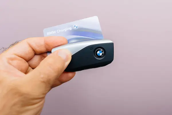 stock image Munich, Germany - Sep 2, 2024: POV of a male hand holding both the BMW charging card and the BMW electric vehicle car key against a pink background, with the key featuring buttons to unlock the car