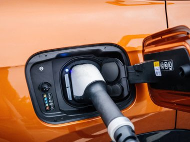 Turuncu lüks elektrikli bir araba hızlı şarj cihazına bağlıdır. Parlayan mavi bir ışık aracın aktif olarak şarj olduğunu ve modern EV teknolojisi sergilediğini gösterir..