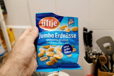 Paris, Fransa - 19 Temmuz 2020: Arka planda parlak mavi ambalaj ve mutfak malzemelerinin yer aldığı bir paket kızarmış ve tuzlu fıstıklı Ultje Jumbo Erdnusse taşıyan bir erkeğin yakın çekimi.
