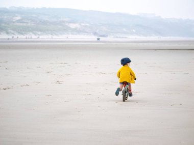 A young child in a yellow jacket rides a small bike on an expansive, empty beach. The scene captures a peaceful moment of outdoor fun and exploration clipart