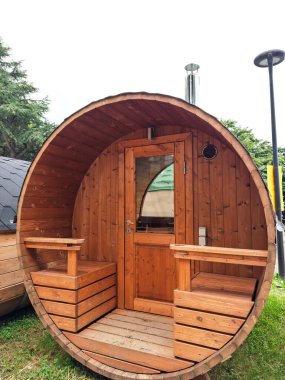 Bahçede, cam kapılı ve banklı yuvarlak bir tasarımı olan ahşap varil şeklindeki bir sauna, rahatlatıcı, kırsal bir refah alanı yaratıyor..