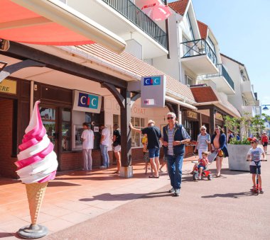 Neufchatel-Hardelot, France - Aug 20, 2024: A lively street where people walk, children play, and a large ice cream cone sculpture stands in front of shops and cafes. clipart