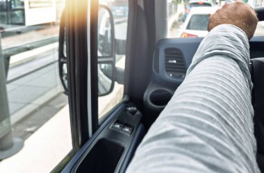 Ağır bir aracın içindeki sürücü görüntüsü yan aynayı ve kollarını kapıda dinlenirken gösterir, trafikte beklerken bir anı yakar - pencereden bir ışık parlaması