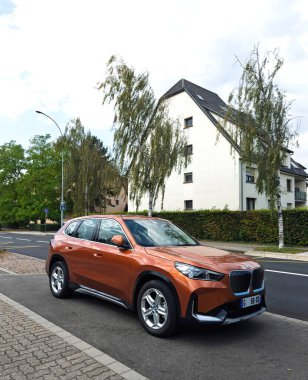 Paris, Fransa - 18 Eylül 2024: Parlak turuncu bir BMW iX1 elektrikli araba sessiz bir banliyö caddesinde park edilmiş, ağaçlar ve evlerle çevrili, şehir içi ulaşımı ve modern araç tasarımını yansıtan