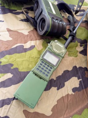 Paris, France - Sep 11, 2024: A green military radio and black binoculars are placed on a camouflage cloth for tactical use - Harris Falcon II clipart