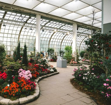 An indoor botanical garden with colorful flower beds, lush greenery, and a glass-paneled roof. Visitors admire plants along the curving pathway on a bright day clipart