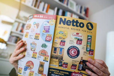 Frankfurt, Germany - Nov 3, 2024: Person reading a Netto supermarket flyer showcasing special discounts on various grocery items, including dairy, snacks, and beverages, against a home background clipart