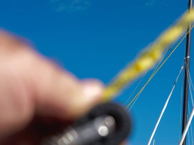 Yakın plan bulanık bir el yelken donanımı halat ve makara ile donatılmış, açık mavi gökyüzü arka planına karşı bir direğe uzanan halatlar üzerine odaklanmış.