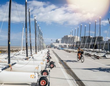 Hardelot, France - Aug 21, 2023: A lineup of land yachts parked on Hardelot Beach, France, with masts and wheels ready for land sailing. Cyclist passing by under a blue sky with scattered clouds clipart