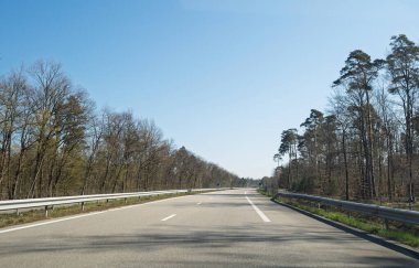 Açık mavi gökyüzünün altında ağaçlarla çevrili geniş, boş bir otoyol. Yolun sağ tarafında yeşil bir işaret var. Huzurlu kırsal alan