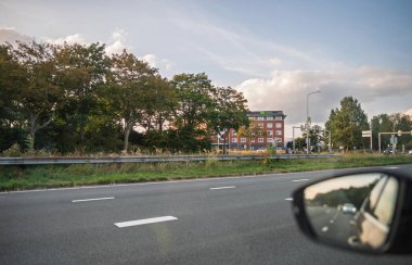 Haarlem, Hollanda - 30 Ağustos 2024: Haarlem, Hollanda 'da yol kenarı manzarası, Ibis oteli, yeşil ağaçlar, hareket eden arabalar ve parçalı bulutlu bir gökyüzüne karşı bir araba aynasında yansıması