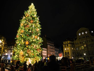 Strasbourg, Fransa - 23 Aralık 2024: Strasbourg şehir merkezinde gece boyunca tarihi binalar ve ziyaretçilerle çevrili büyük, süslü bir Noel ağacı parlıyor