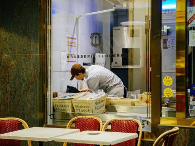 Strasbourg, France - Dec 23, 2024: Chef preparing food in the kitchen of Brasserie Flo in Strasbourg, viewed through a window with oyster boxes and cozy bistro seating outside clipart