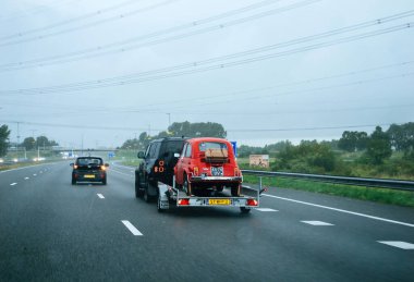 Den Helder, Netherlands - Aug 24, 2024: A classic red Fiat car on a trailer, seen on a Dutch highway during rainy weather, showcasing vintage automotive transport in Europe clipart