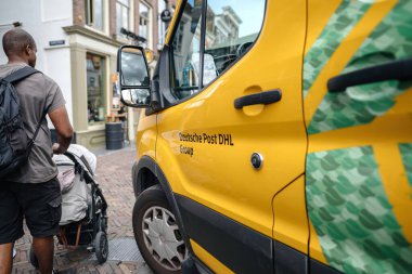 Alkmaar, Netherlands - Aug 30, 2024: A Black man pushes a stroller past a Deutsche Post DHL yellow van in the city center of Alkmaar, Netherlands, capturing a moment of urban life and diversity clipart