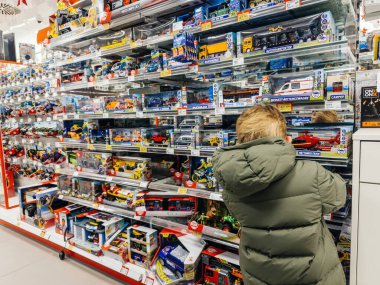 Berlin, Almanya - 11 Aralık 2024: Yeşil ceketli bir çocuk, çeşitli renkli seçenekler sunan bir dizi oyuncak araç ve set ile dolu bir oyuncak mağazası koridorunu inceliyor.