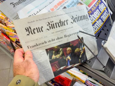 Berlin, Almanya - 6 Aralık 2024: Neue Zuricher Zeitung gazetesini bir gazete bayisinde tutan ve tanınmış uluslararası manşetlerin yer aldığı ve renkli dergilerin kapaklarıyla çevrili bir kişi.