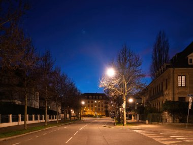 Alacakaranlıkta Strasbourg 'daki Rue de Verdun' un sakin manzarasında çıplak ağaçlar, aydınlatılmış sokak lambaları ve derin mavi gökyüzüne karşı zarif bir mimari yer alır.