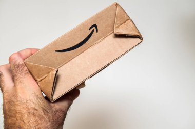 Paris, Fransa - Bölüm 22, 2021: Amazon gülümseme logosunun yer aldığı küçük bir karton kutuyu kavrayan bir el, tarafsız bir arkaplana karşı yakın plan perspektifinde sergilendi.